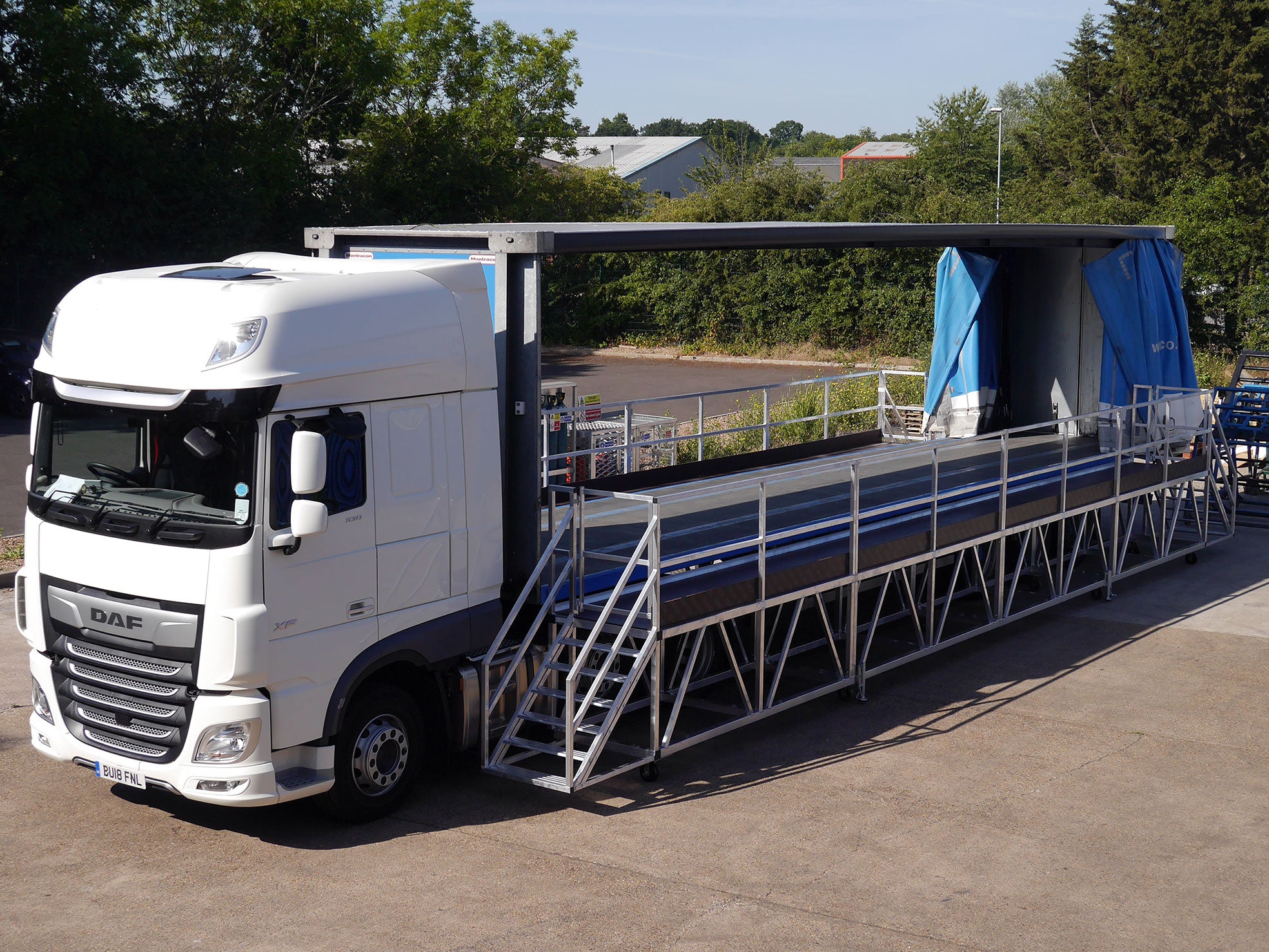 White truck with a large mobile platform structure on a paved area with trees in the background